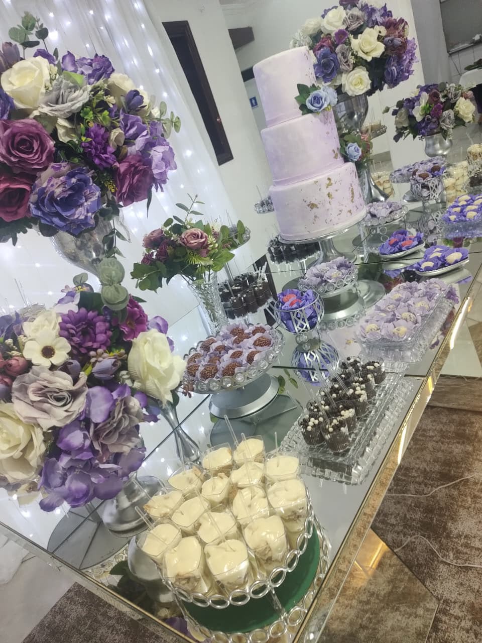 Mesa de doces com bolo, arranjos de flores em tons de roxo e branco e doces finos