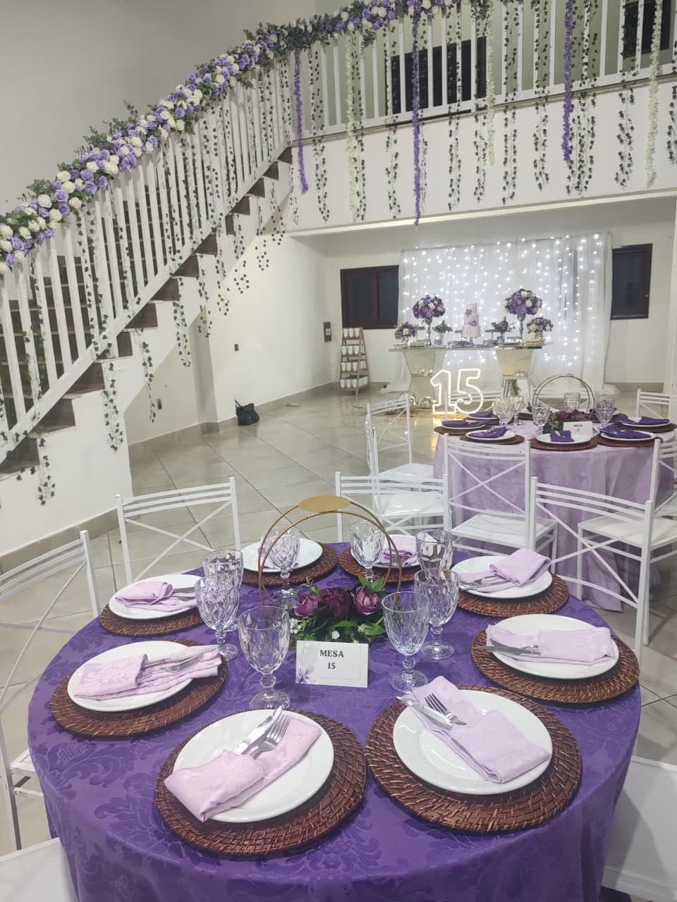 Sala de eventos decorada com mesa de festa, escada com flores, iluminação de led e painel de doces