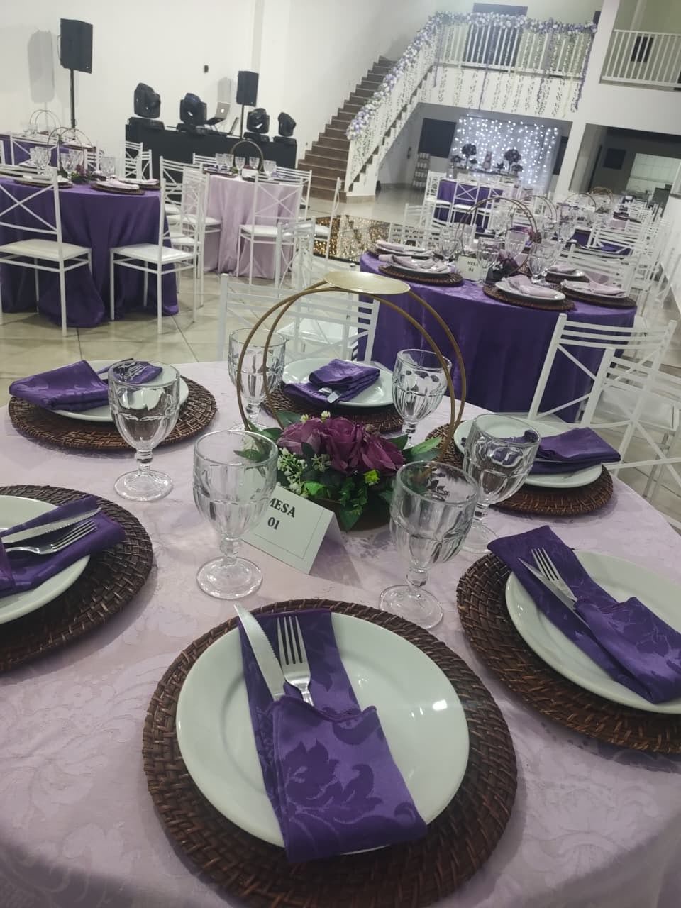 decoração de festa ou casamento com mesas redondas com toalhas lilás e roxas, cadeiras brancas, arranjos de flores roxas e placas indicando as mesas num salão com escada ao fundo