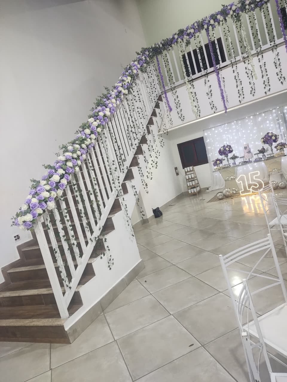 Interior de um salão de eventos com escada decorada em cores lilás e branco, toalhas e decoração para uma festa de quinze anos