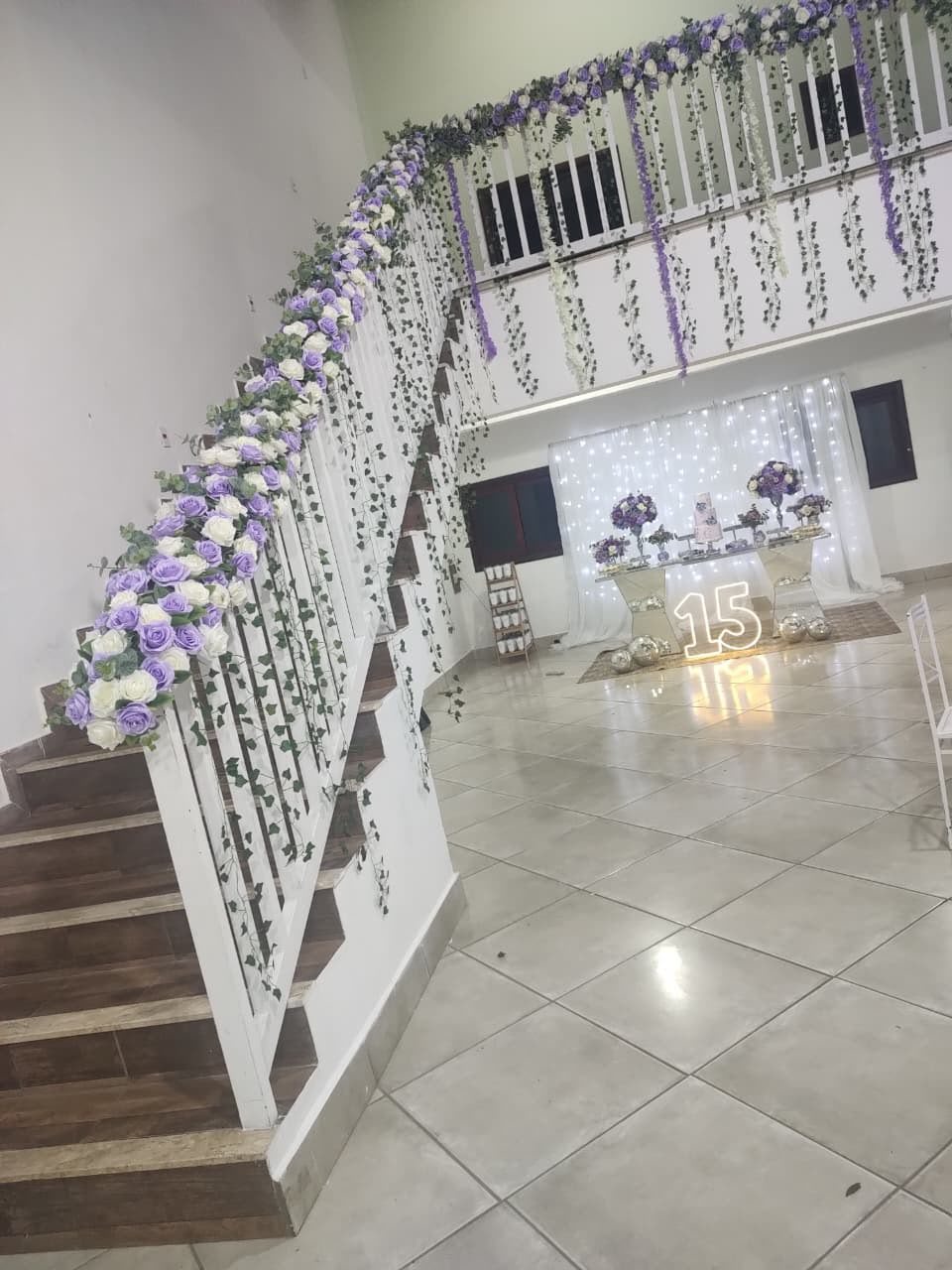 Ambiente de evento decorado com flores na escada, num espaço com piso de tile, balcão com decoração e número 15