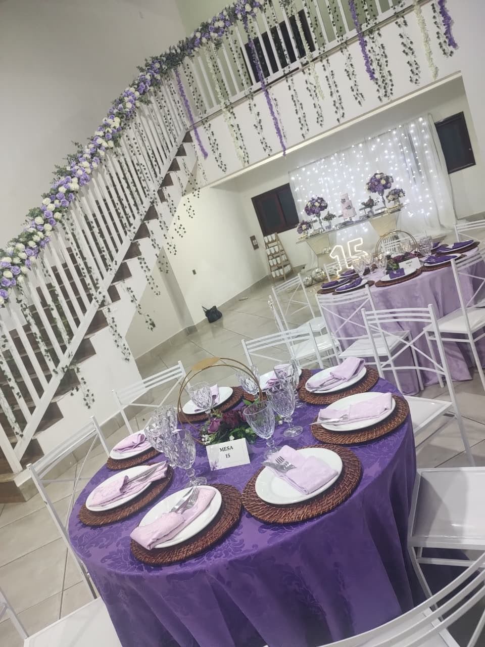 decoração de festa de 15 anos com escada enfeitada de flores roxas, mesa redonda com toalha lilás e fundo iluminado