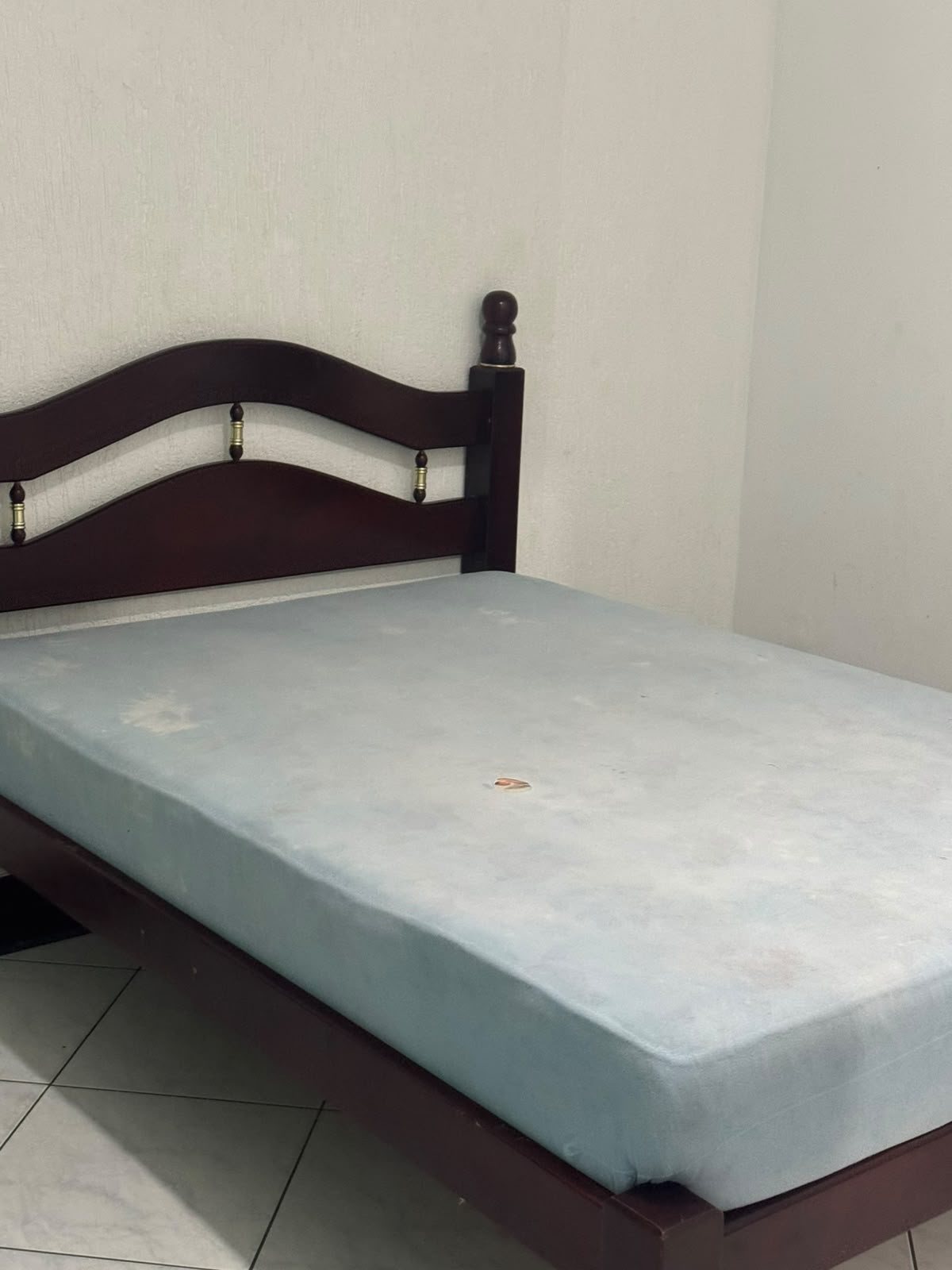 uma cama de madeira com cabeceira curva e colchão de cor clara numa sala com piso de azulejo e parede em branco  — texto: