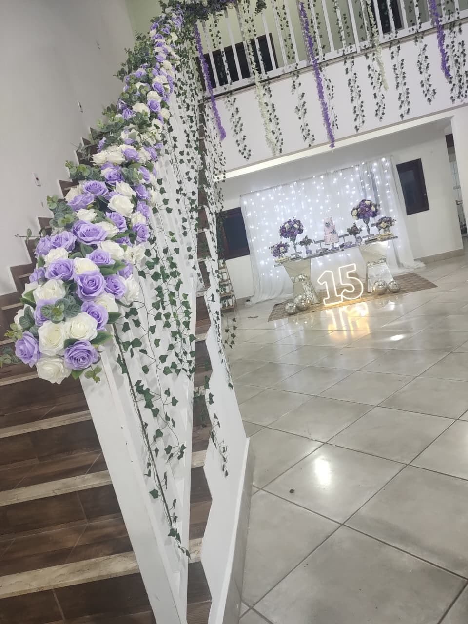 Ambiente de evento decorado com flores lilás e brancas no balcão de escada, tablete com decoração e o número 15 iluminado no chão